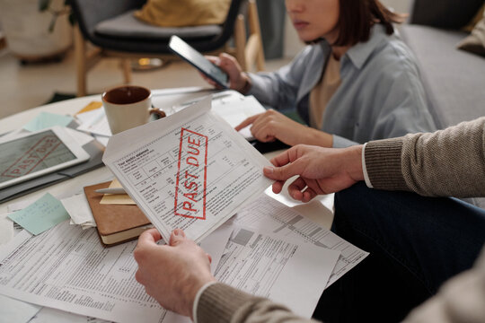 Unpaid financial bill with past due stamp in hands of young man sitting by table with documents against his wife making calculations