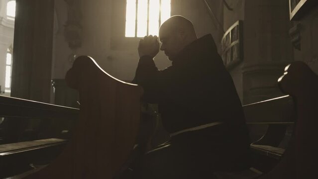 Mid adult caucasian Christian monk in robe praying in church side view medium shot slow motion. Religious man sitting on cathedral pew bench with clasped hands and looking up. Worship concept