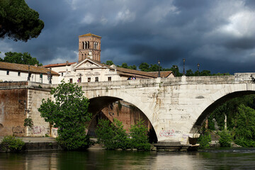 Fototapeta premium Ponte Fabricio in Rome which connects with the Tiber Island, bell tower of the Basilica of San Bartolomeo in the background, dark sky full of clouds.