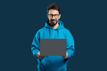 A man wearing a blue hoodie is standing while holding a laptop in his hands. He appears focused on...