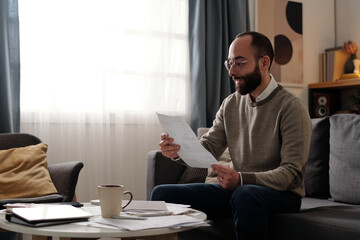 Smiling bearded man with financial paper document looking through data while sitting on couch in...