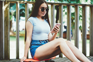 Attractive teenage hipster girl wear in sunglasses sitting on mini board while communicated via smartphone, beautiful woman searching information using mobile phone connected to 4g internet in roaming