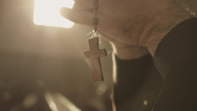 Christian rosary with cross hanging from male hands and swinging close up. Monk holding prayer beads in clasped arms while praying in cathedral closeup. Christianity concept