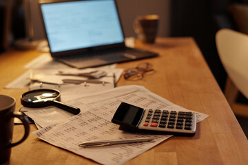 Calculator, silver pen and magnifying glass on financial bills containing unpaid sums lying on table with laptop on background