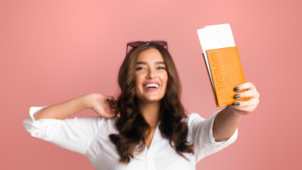 A young woman with wavy hair and sunglasses pushed up on her head is joyfully showing passport with...