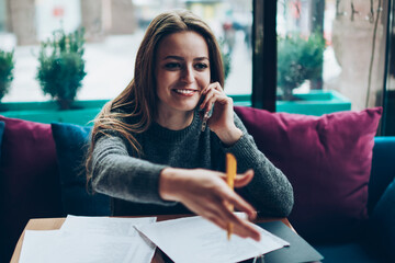 Positive smiling female economist calling friend using application on modern mobile phone in work...