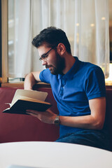 Handsome calm hipster guy reading interesting book while resting at cozy cafeteria, young concentrated male student in stylish spectacles relaxing and enjoying of romantic novel indoors