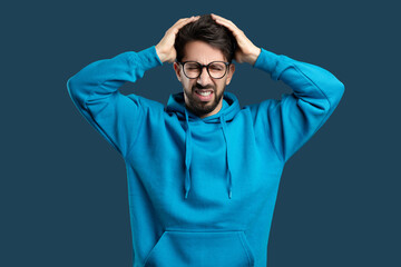 A young man with a beard and glasses, wearing a blue hoodie, is standing against a solid blue...