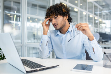 A young adult male appears stressed and tired while working on his laptop in a bright modern office, with a smartphone on the desk next to him.