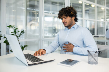 Fototapeta premium A young adult male seated at his office desk shows signs of pain, clutching his stomach while looking distressed, indicating discomfort or health issues during work.
