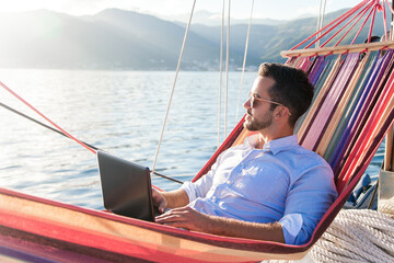 Freelancer working in traveling, using laptop, Internet. Man with computer in beach hammock. Sea...