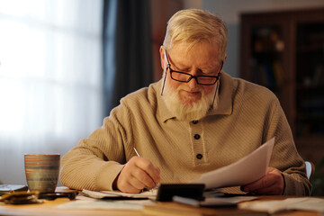 Bearded senior man in eyeglasses bending over table and making notes in notepad while checking information in financial bills