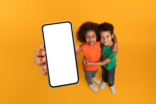 African American kids holding up a cell phone with a blank white screen, showing no content or images, mockup copy space, yellow studio background