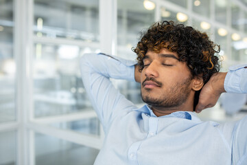 Obraz premium A young man in a light blue button-down shirt relaxes with eyes closed, stretching his neck while seated in a bright modern office space, displaying feelings of ease and peace.