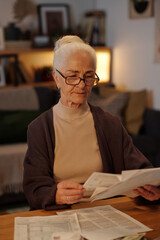 Aged woman in eyeglasses and casual attire sitting by table in home environment and looking through financial data in bills