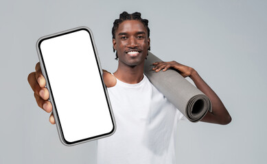 Athletic black man is holding fitness mat and a cell phone with white blank screen in his hand,...