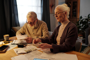 Senior woman with white hair sitting by table and counting dollar bills over paid and unpaid financial bills against her husband making notes