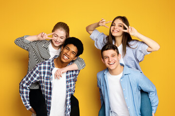 A cheerful group of four young friends is playfully posing together, with two making peace signs near their eyes and all sporting genuine, joyful smiles.