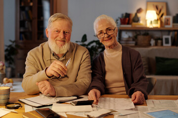 Smiling senior couple in casual attire looking at camera while sitting by table with housing payment bills and checking information