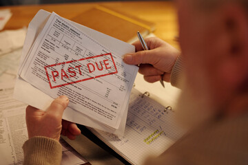 Hands of aged man holding silver pen, envelope and unpaid financial bill with past due stamp while sitting by table and checking post