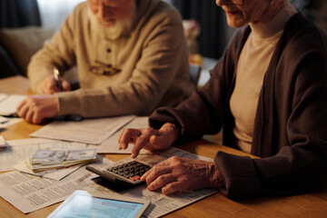 Cropped shot of senior woman in casual apparel sitting by table with financial documents and using calculator against her husband