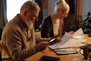 Side view of aged man with magnifying glass looking through financial bill while his wife sitting next to him and checking data in documents
