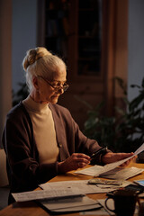Slim aged woman with white hair sitting by table in home environment and looking through housing payment bill with magnifying glass