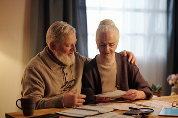 Senior man with grey beard supporting his wife with post due bill looking through information and expressing worry and anxiety