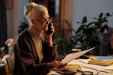Senior woman with white hair sitting by table with financial bills and speaking to worker of housing service on mobile phone