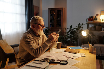 Side view of senior bearded man in eyeglasses using calculator in smartphone while sitting by table with housing payment bills