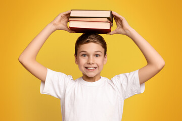 Positive schoolboy holding stack of books on his head, orange studio background