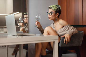 a beautiful girl with short hair and glasses is sitting indoors at a laptop, with her feet on the...