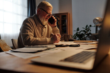 Serious senior man in eyeglasses looking through financial bill while sitting by table at home and...