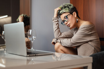 a beautiful girl with short hair and glasses is sitting indoors at a laptop chatting and working...