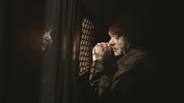 Christian monk in robe praying and asking for forgiveness in church confessional booth. Mid adult caucasian religious man sitting near confession window during prayer side view