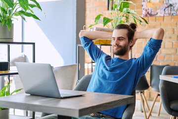 Satisfied relaxed young man looking at laptop screen while sitting in coworking cafe