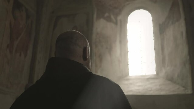 Bald caucasian Christian monk wearing monastery robe walking and looking out of sunlit window in church back view medium shot. Devotion and faith concept