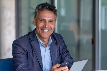 Fototapeta premium Middle aged Hispanic business manager ceo using cell phone mobile app, laptop. Smiling Latin or Indian mature man businessman holding smartphone 