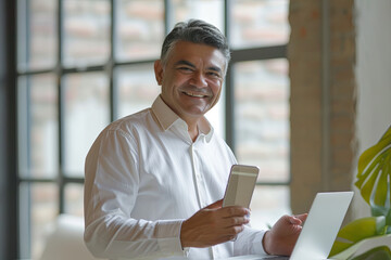 Middle aged Hispanic business manager ceo using cell phone mobile app, laptop. Smiling Latin or Indian mature man businessman holding smartphone
