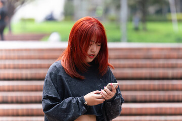 Korean woman with bright red hair absorbed in texting on her smartphone