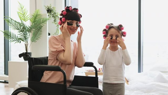 Young woman and cute daughter doing hair perm and applying patches. Happy Caucasian mother wheelchair user and little daughter doing cosmetic procedures at home in bedroom.