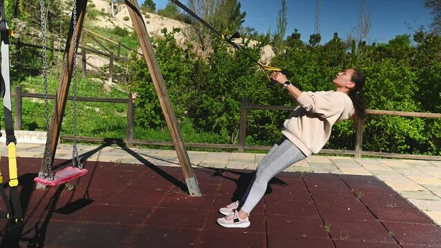 Full length side portrait of a middle aged sporty woman 40s, doing pulls up exercises on arms with suspension straps, working out on the urban sports ground in the nature outdoors . People and fitness