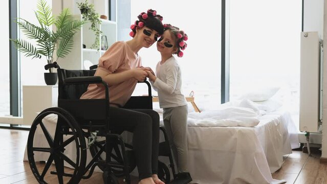 Happy Caucasian mother wheelchair user and little daughter doing cosmetic procedures at home in bedroom. Young woman and cute daughter doing hair perm and applying patches.