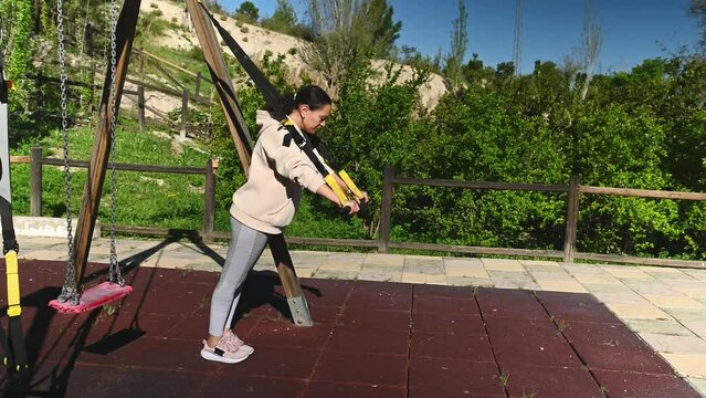 Full length side portrait of a middle aged athletic woman 40s, doing push up arms exercises with suspension straps, working out on the urban sports ground in the nature outdoors . People and fitness