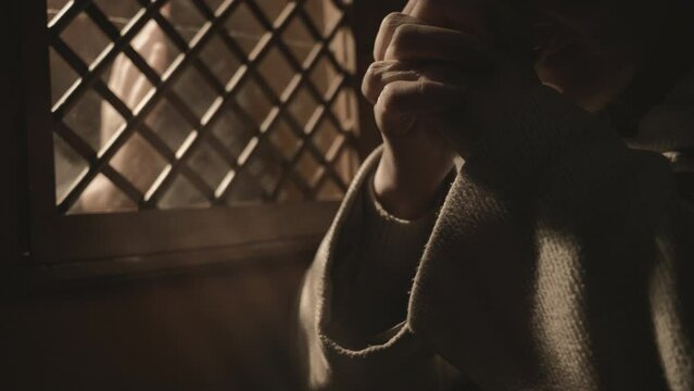 Mid adult religious woman praying near confessional booth window. Female parishioner sitting with clasped hands during confession in church close up