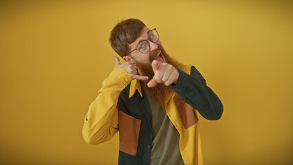 Young redhead bearded man making call me gesture and pointing at camera isolated over yellow...