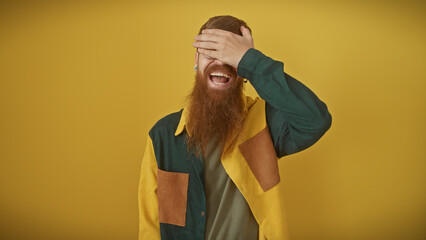 A cheerful, young, redhead man with a beard, laughing while covering his eyes, stands against an...