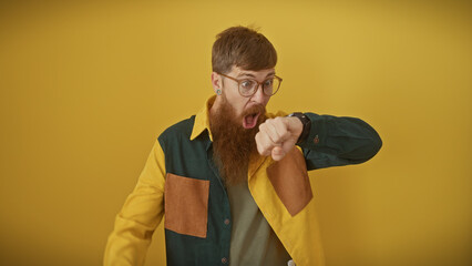 A surprised redhead man with beard and glasses checks the time on his watch against a yellow...
