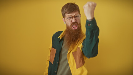 Angry redhead bearded man raising fist against a yellow background, portraying frustration.