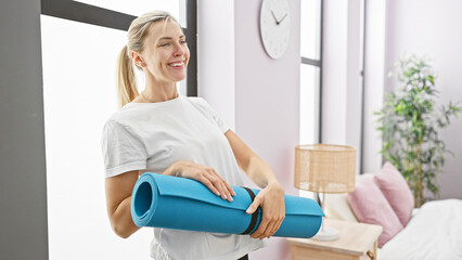 A smiling caucasian woman holds a yoga mat in a bright bedroom, illustrating wellness and a healthy...